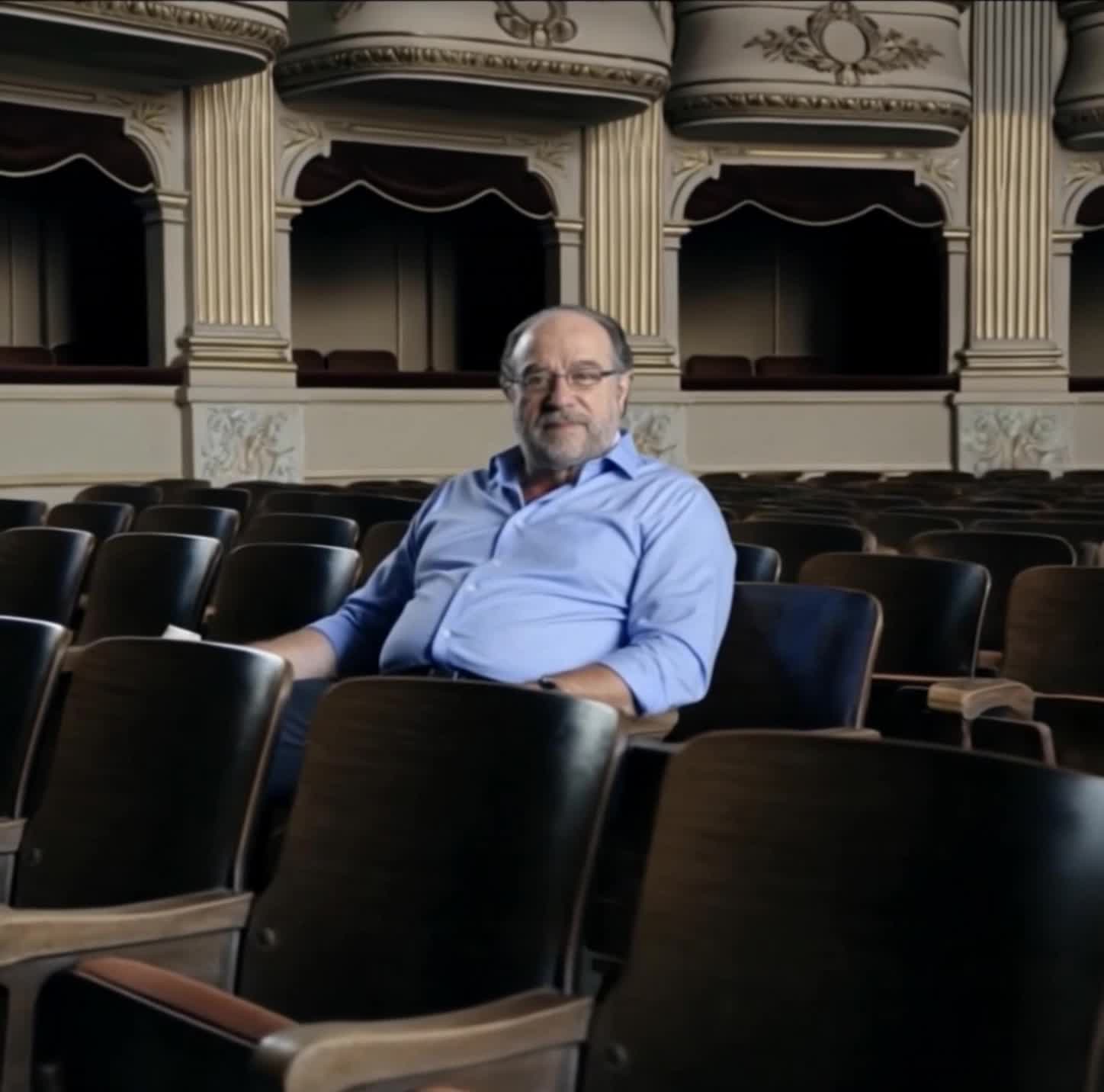 The camera slowly pulls back from the man, gradually revealing that he is seated in a grand theatre 