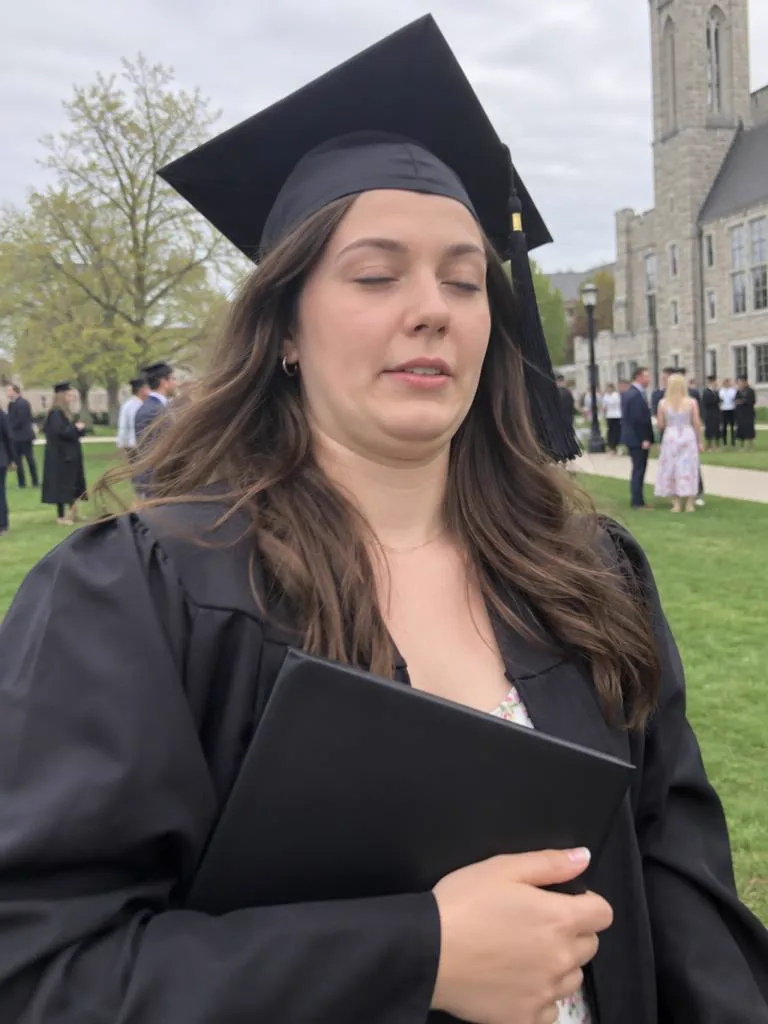 Take 1: a graduate in a black cap and gown holding her diploma, eyes closed mid-blink